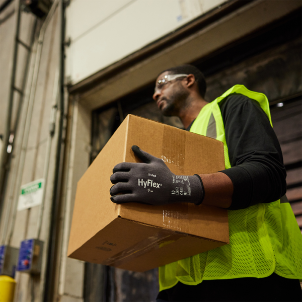 Worker wearing HyFlex 11-840 gloves lifting a cardboard box in a distribution warehouse.