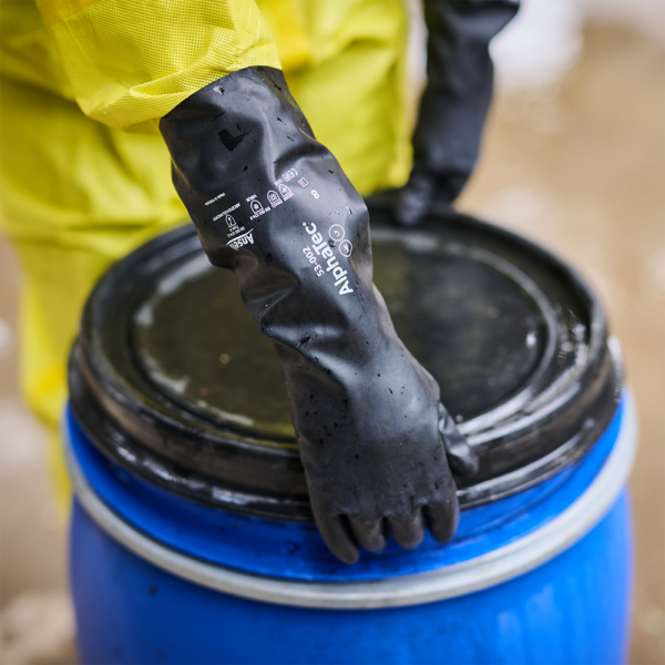 Worker wearing Ansell AlphaTec 53-002 gloves opening black drum lid in spill-prone area.
