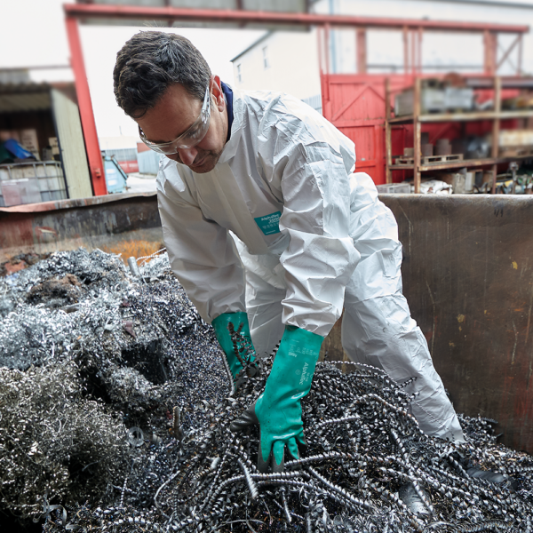 Worker wearing Ansell AlphaTec 58-735 gloves and white chemical coveralls handling metal shavings in an industrial setting.