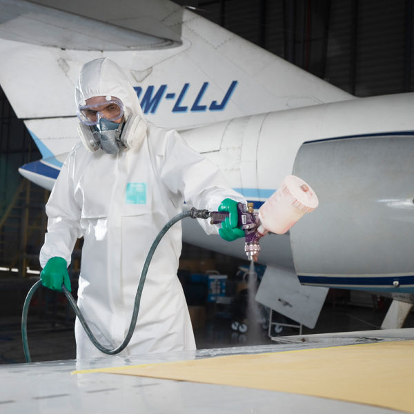 Worker in full PPE spraying coating onto an aircraft surface, wearing chemical-resistant gloves and a respirator