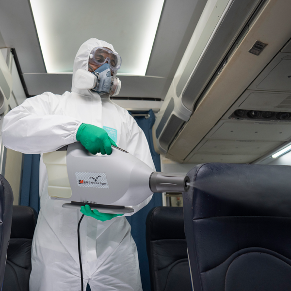 Technician in full-body coveralls and gloves disinfecting an aircraft cabin using a fogging machine