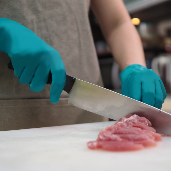 Worker wearing teal chemical-resistant gloves while slicing raw meat on a cutting board in a commercial kitchen