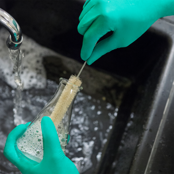 Gloved hands scrubbing a laboratory flask under running water using a bottle brush