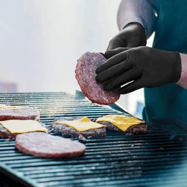 Person wearing Ansell MICROFLEX 93-732 black nitrile gloves grilling burgers and handling raw meat safely.