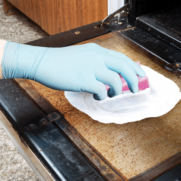 Worker wearing Ansell MICROFLEX 93-833 disposable nitrile gloves scrubbing an oven door surface with a sponge.